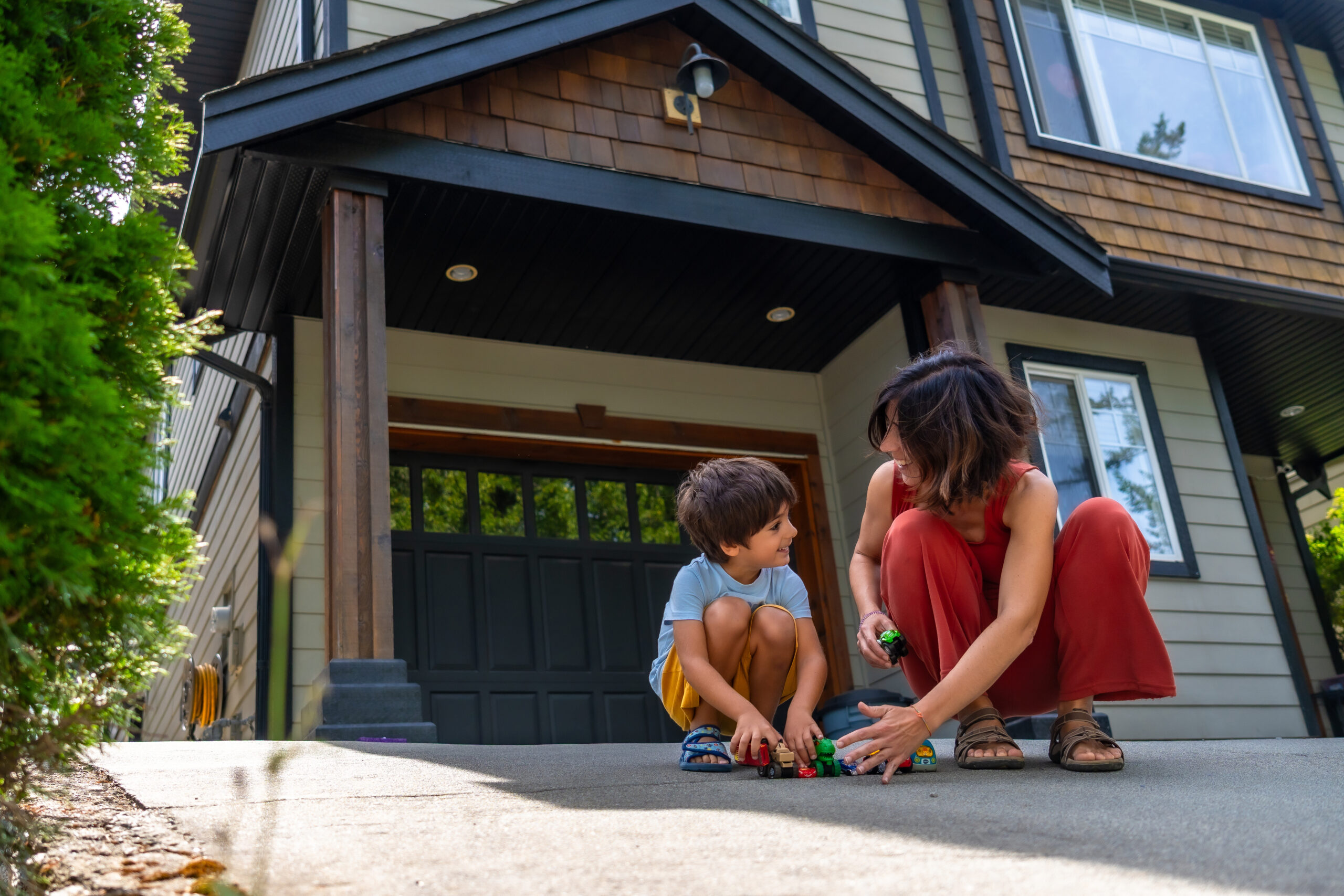 Mother and her young son are happily playing with toy cars on the driveway of their modern two story house, enjoying quality time together on a sunny summer day to demonstrate importance of garage door safety